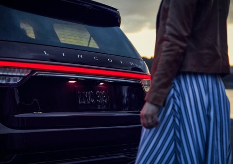 A person is shown near the rear of a 2024 Lincoln Aviator® SUV as the Lincoln Embrace illuminates the rear lights | Klaben Lincoln of Warren in Warren OH
